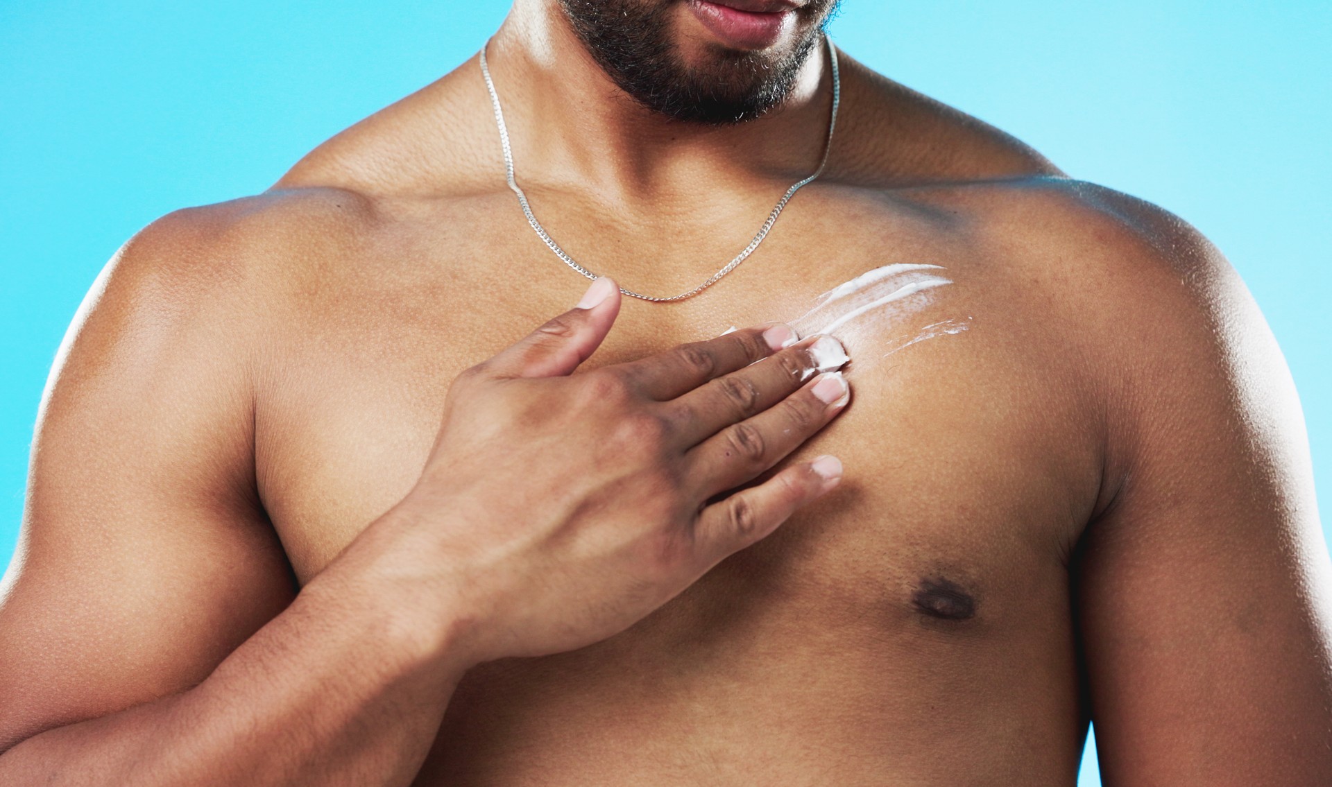 Skincare, applying and man with cream on his chest isolated on a blue background in a studio. Moisturizing, sexy and male model with sunscreen, lotion or moisturizer on his body for grooming